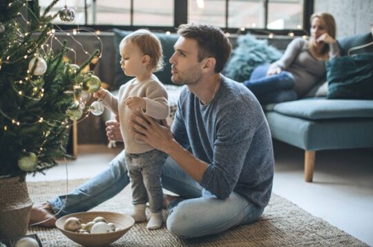 Man with child in front of Christmas tree with a woman looking on.