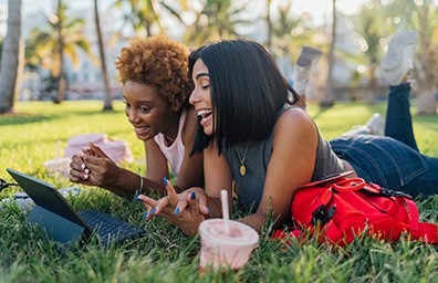 Two young woman laying in grass with smart devices
