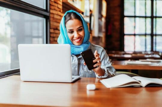 woman at laptop looking at phone.