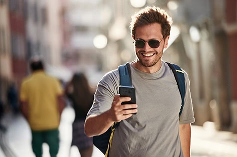 Happy male wearing backpack text messaging on smart phone during spring day on the street.
