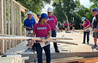 PenAir Team members wearing berry tshirts and blue ice penair logo lift pieces of wood to help build a house.
