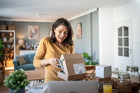 Young woman is happily surprised while unpacking a delivery box in her living room, surrounded by other packages and working on her laptop.