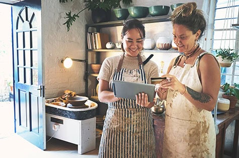 two young women using a digital tablet and credit card while working together in a pottery studio